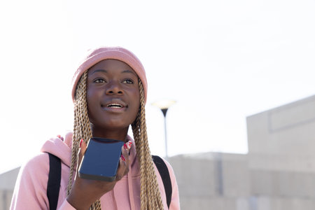 Teen African American girl talking into phone at urban plaza in pink beanie and hoodie, backpack....