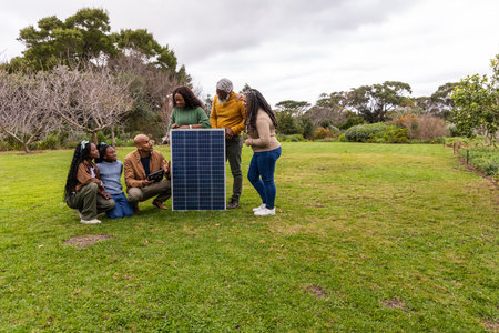 African American family studying solar panel, kneeling man holding tablet in garden, wearing coats....