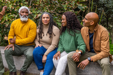 African American friends sitting on wooden log bench at park wearing knit sweaters, holding hands. Quartet, outdoor, greenery, woodland, casual, cozy, conversationの写真素材