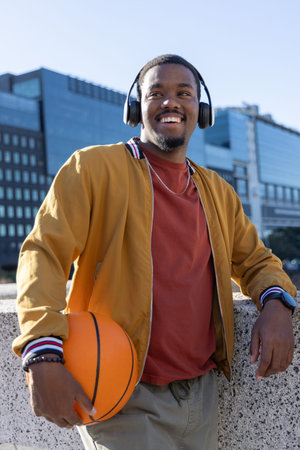 African American man leaning on rail, holding orange basketball, wearing headphones. Promenade, urban, bomber, casual, ball, smartwatchの写真素材