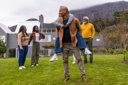 African American family wearing jackets standing on lawn by home while man carrying teenager....