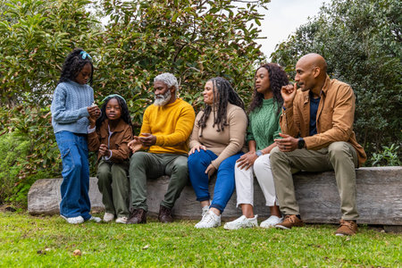 African American family with kids and seniors sitting on log in park sharing smartphone in sweaters....