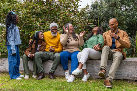 African American family in knit sweaters laughing, sitting on wooden log bench on lawn holding cup....
