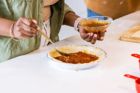 African American woman in green jacket and companion spreading jam into aluminum pie tin on counter. Kitchen, baking, duo, homemade, spread, preserve, fruitの写真素材