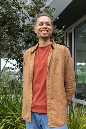 Man in his 30s standing in courtyard near glass panels, wearing rust T-shirt, among tall grasses....