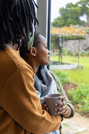 African American couple standing at large window, woman holding ribbed mug, partner embracing waist....