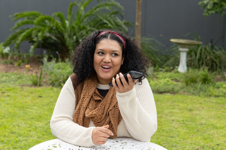 Woman sitting at white garden table holding smartphone near mouth speaking, wearing brown scarf....