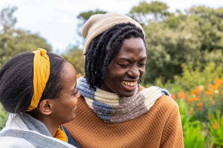 African American couple laughing and embracing in garden wearing knit beanie, striped scarf, shawl....