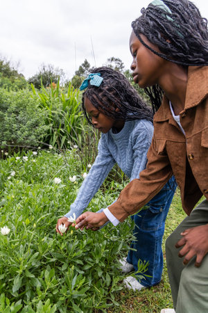 Two African American female children kneeling leaning over foliage in park touching white flowers....