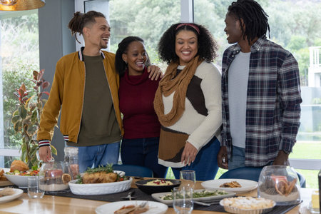 Diverse friends standing together behind dining table at home, sharing roasted chicken and sides....