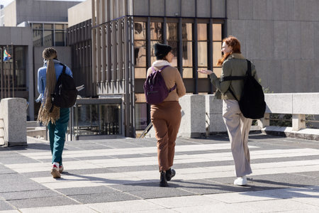 Three women walking across university plaza, talking and gesturing with backpacks and beanie....