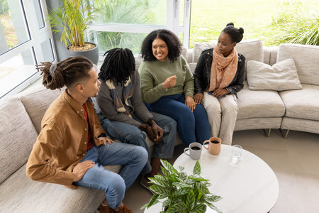 African American woman in green sweater and jeans smiling, gesturing in living room by coffee table....
