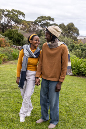 African American couple walking hand-in-hand on manicured lawn in garden wearing knit scarves. Partners, romance, stroll, park, autumn, outdoors, natureの写真素材
