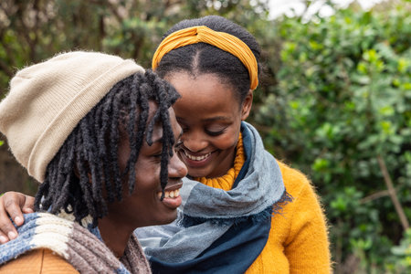 African American couple standing in garden, smiling and leaning, wearing beanie, headband, scarves....