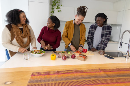 Diverse friends chopping vegetables and tossing salad on white kitchen island with cutting board....
