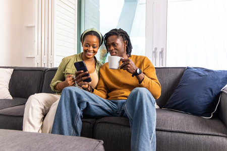 African American couple sitting on dark gray sofa in living room, sharing smartphone and mug. Duo, phone, screen, home, sunlit, cozy, modernの写真素材
