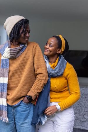African American couple standing close, smiling, wearing knit scarves and beanie in living room....