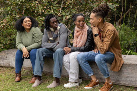 Group of four friends chatting and sitting on wooden log bench in public garden wearing scarves....