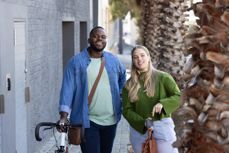 Diverse couple walking together along narrow tree-lined urban sidewalk, man holding bicycle handles....