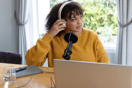 Adult woman adjusting headphones preparing to speak into mic at wooden table in mustard sweater....