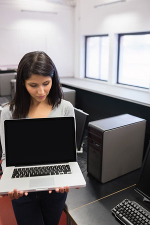 Female wearing light gray long-sleeve top holding open laptop with black screen in computer lab....