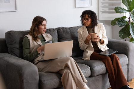 Diverse women colleagues sitting on sofa in lounge, using laptop, tablet and mugs in blazer, vest. Calm, professional, collaboration, discussion, interior, workspace, devicesの写真素材
