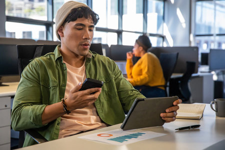 Diverse coworkers in office Asian man wearing beanie holding phone using tablet viewing chart....