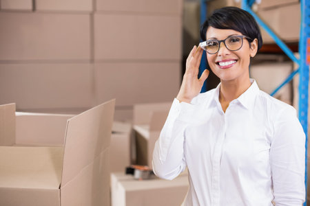 Mature woman white top holding specs standing in depot near stacked boxes tape visible, copy space. Warehouse, logistics, supervisor, packaging, shelving, stockroom, inventoryの写真素材