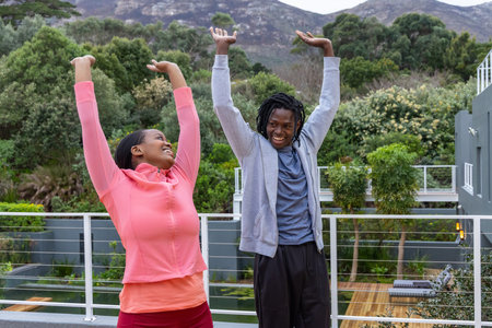 African American couple stretching in athletic clothes on balcony with metal railing and mountains....