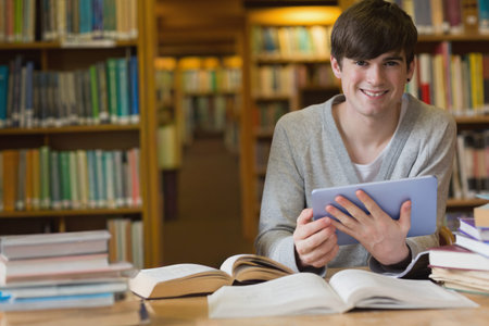 Adult male student studying at study table holding tablet with open books and bookshelf, copy space....