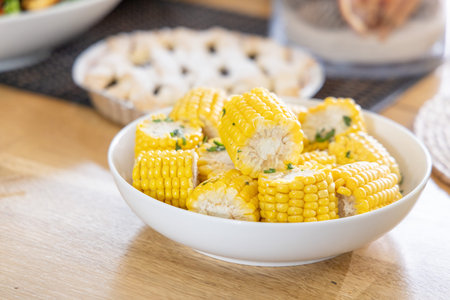 White ceramic bowl holding buttered corn rounds with chopped parsley on light wooden dining table....
