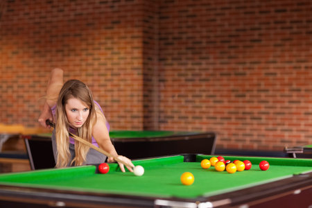 Woman aiming shot at billiards hall leaning over pool table holding cue stick by cue ball. Player,...