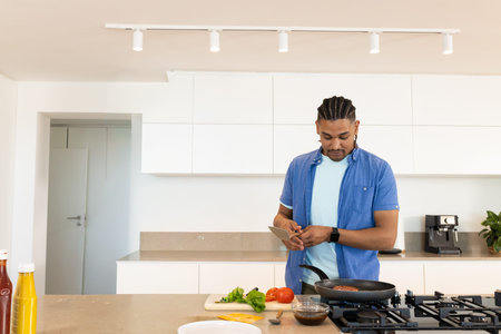 Man in shirt cooking on kitchen island, checking phone, frying pan heating on cooktop, copy space. Modern, interior, timer, tablet, recipe, culinary, brightの写真素材