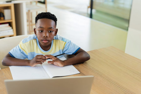 African American child sitting at home study table holding pen writing in notebook near laptop....