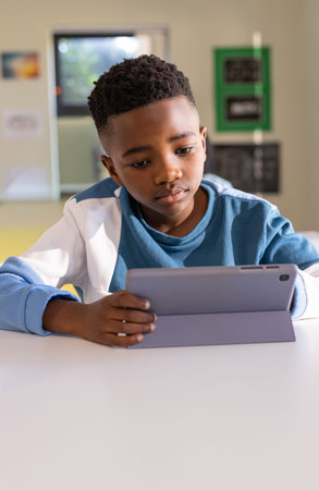 African American child wearing blue crewneck jacket draped sitting in classroom using tablet case....