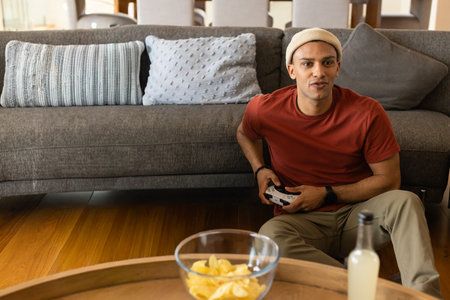 Adult man in beanie sitting in living-room holding controller, chips and drink on table, copy space....