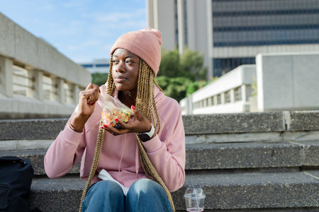 African American woman sitting at campus steps wearing pink hoodie, eating salad from clear box. Urban, outdoors, plaza, casual, student, backpack, lunchの写真素材