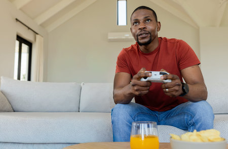 African American man leaning forward playing with white gamepad on sofa by coffee table. Couch, gaming, controller, home, daylight, cozy, relaxationの写真素材