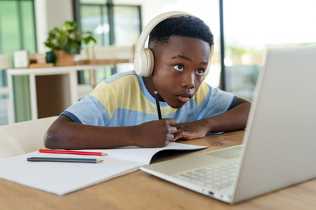 School-age African American boy sitting at home studying on laptop with pencil and headphones....