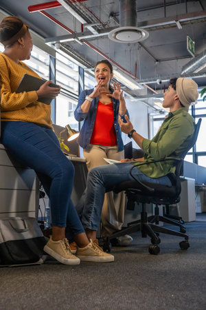Diverse team discussing work by big windows in open office in jeans, holding tablet and papers....