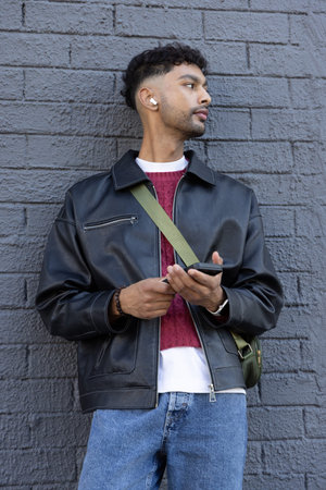 Indian man standing by gray brick wall, holding smartphone, wearing earbud and leather jacket....