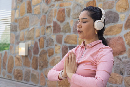 Midadult woman standing on patio by stone wall wearing pink ziptop and white headphones, copy space....