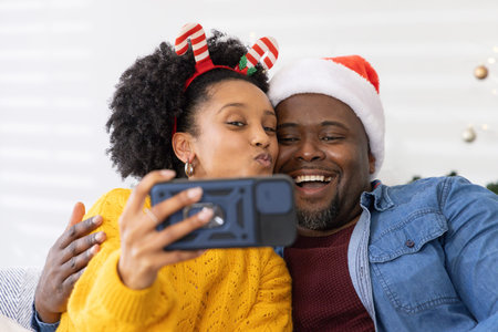 African American couple taking selfie on couch at home with smartphone, Santa hat, headband. Two,...