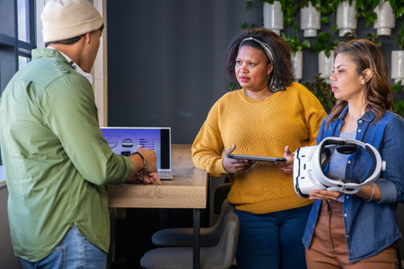 Diverse coworkers leaning around wooden counter in modern office with laptop, tablet, VR headset. Collaboration, teamwork, ideation, design, innovation, workspace, daylightの写真素材