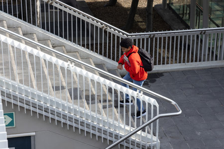 Teen male ascending concrete campus stairs adjusting backpack strap wearing red hoodie, copy space....