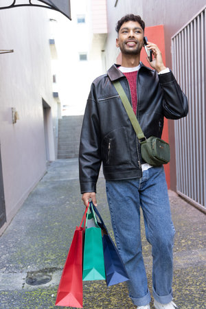 Indian man in twenties walking in narrow alley wearing leather jacket, holding phone, carrying bags....