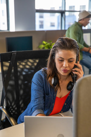 Female worker sitting at desk using laptop and talking on smartphone with headphones in office....