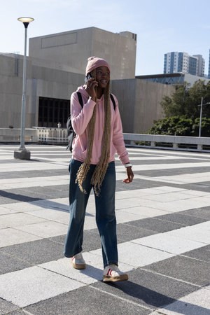 African American female walking and talking on smartphone in plaza wearing pink hoodie and backpack....