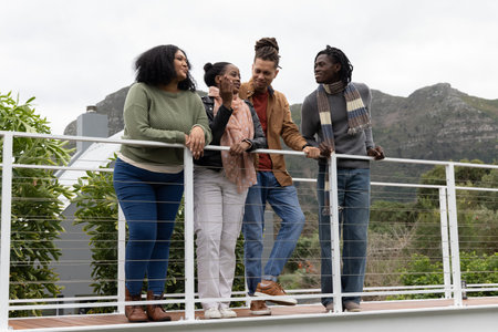 Diverse friends leaning and chatting on raised deck wearing sweaters and scarves by metal railing....