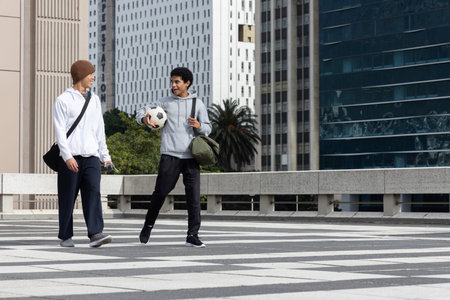 Diverse male friends walking across striped plaza carrying soccer ball, green duffel, tan beanie. Urban, downtown, cityscape, rooftop, highrise, daylight, casualの写真素材